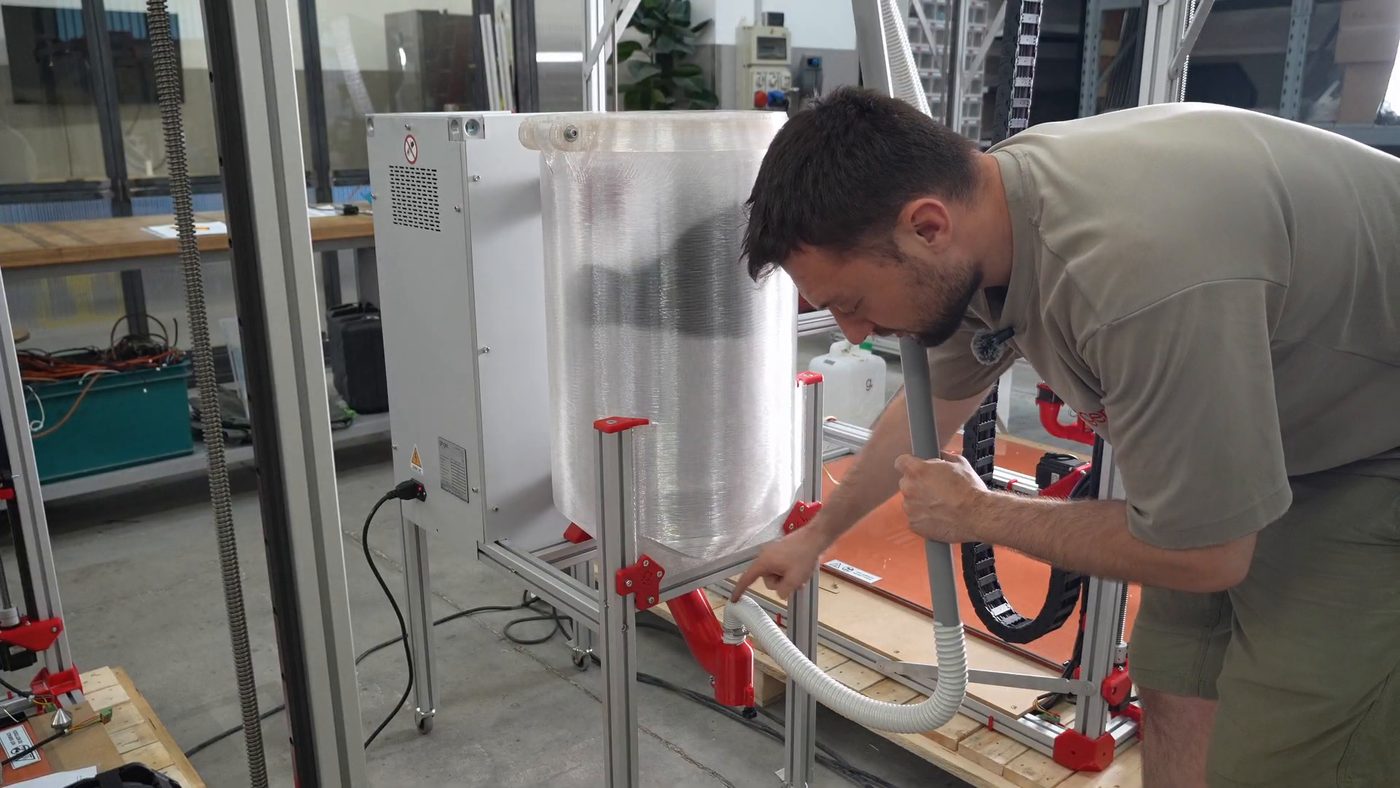The D1 dryer standing beside the printer, its translucent tank on a wheeled frame, with the red pellet gate at the bottom and the white corrugated tube clamped to it