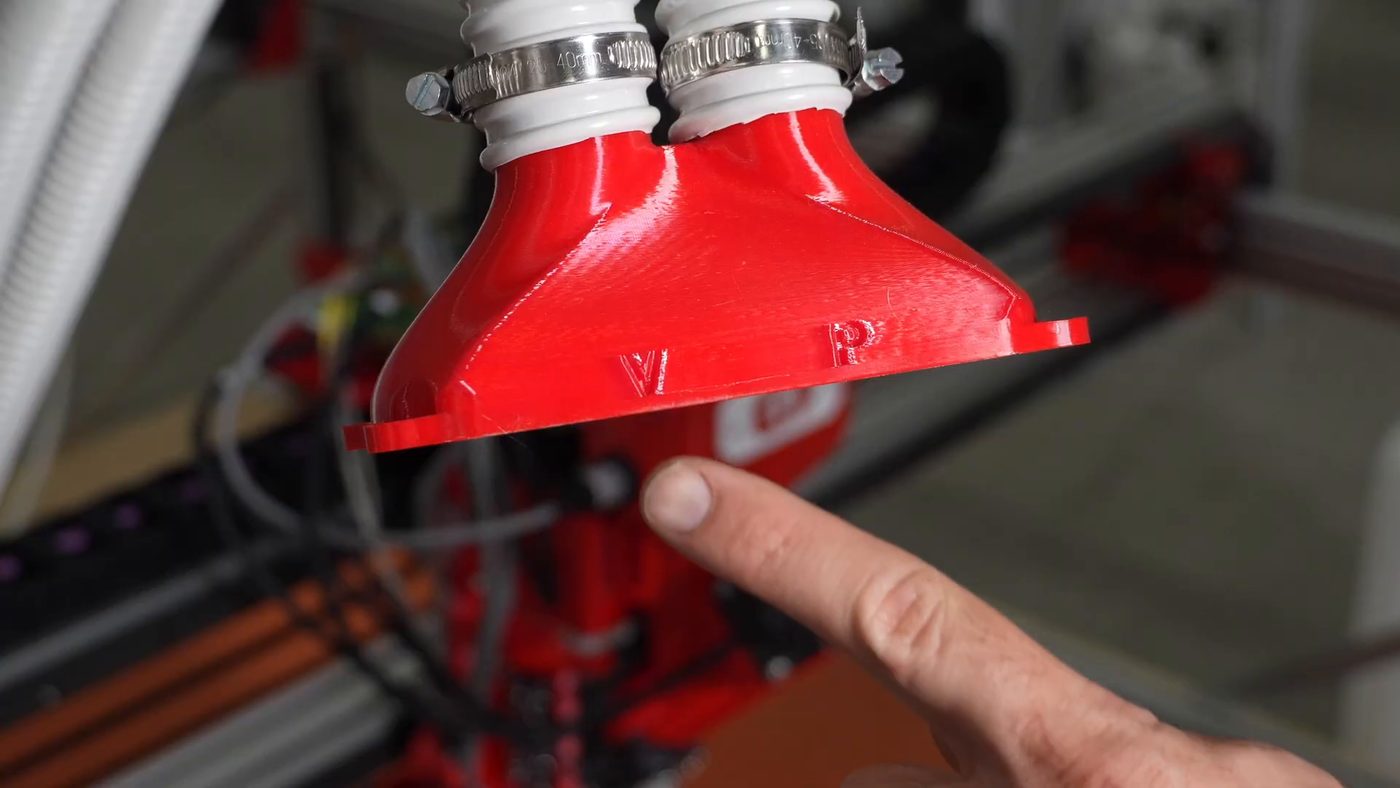 The E17 hopper cap held in one hand, off the printer. It is a red moulded cone with two white tube stubs on top, each secured with a metal hose clamp