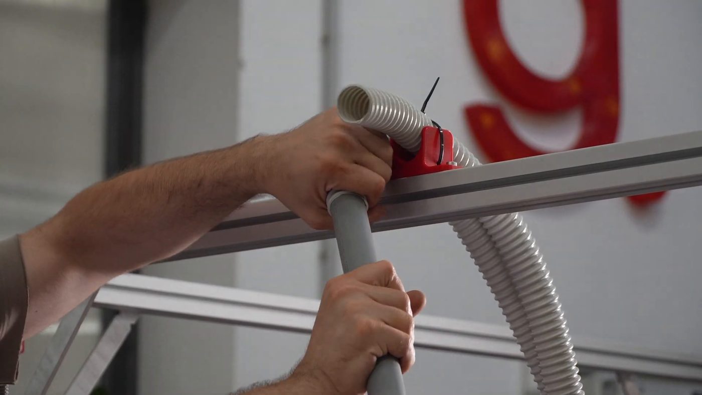 Two hands joining the smooth rigid grey pipe to the white corrugated tube alongside the printer frame