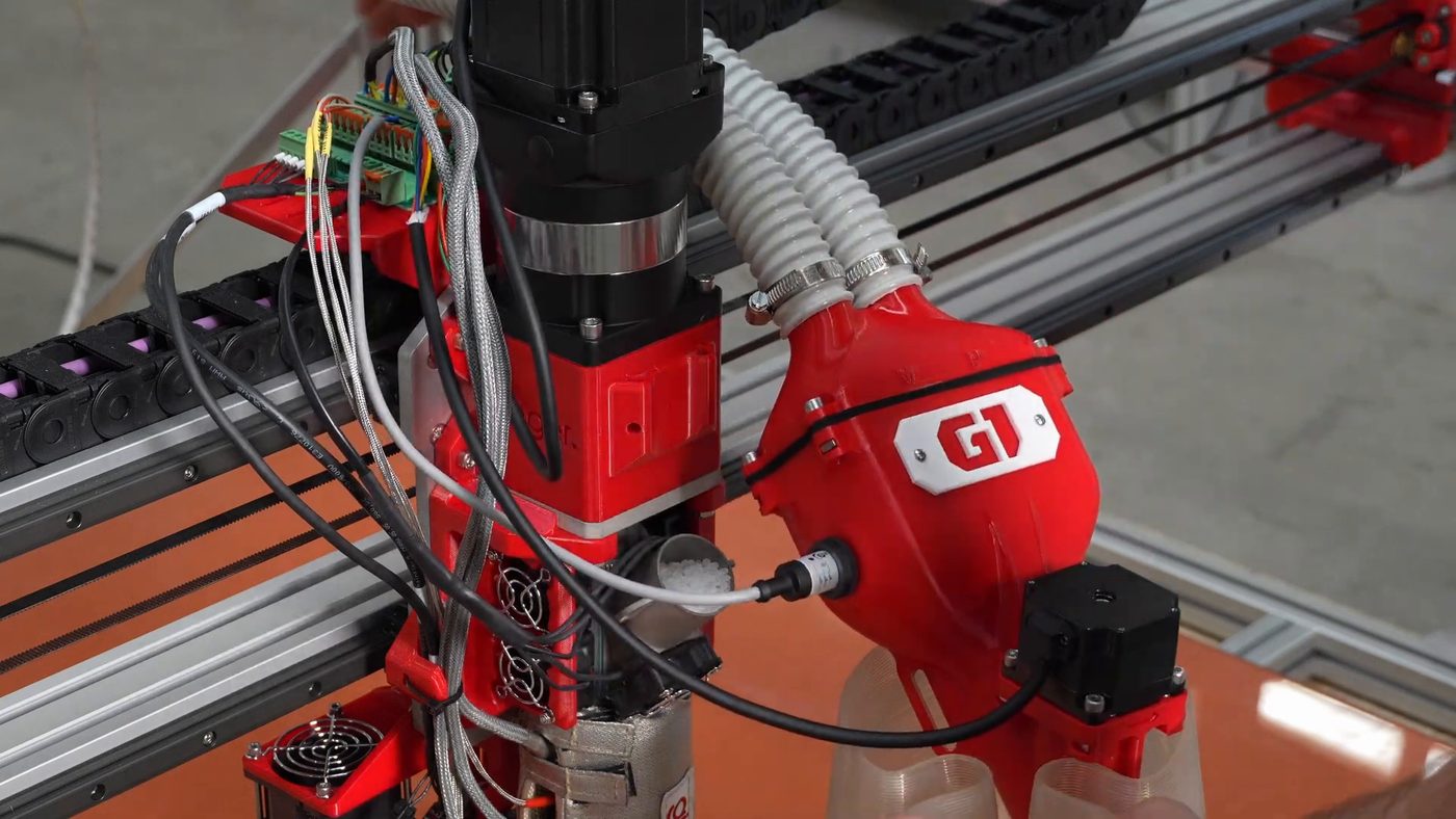 The hopper lifted off the extruder and held tilted over a clear container, pellets running out of its outlet, with the two corrugated tubes still attached at the top