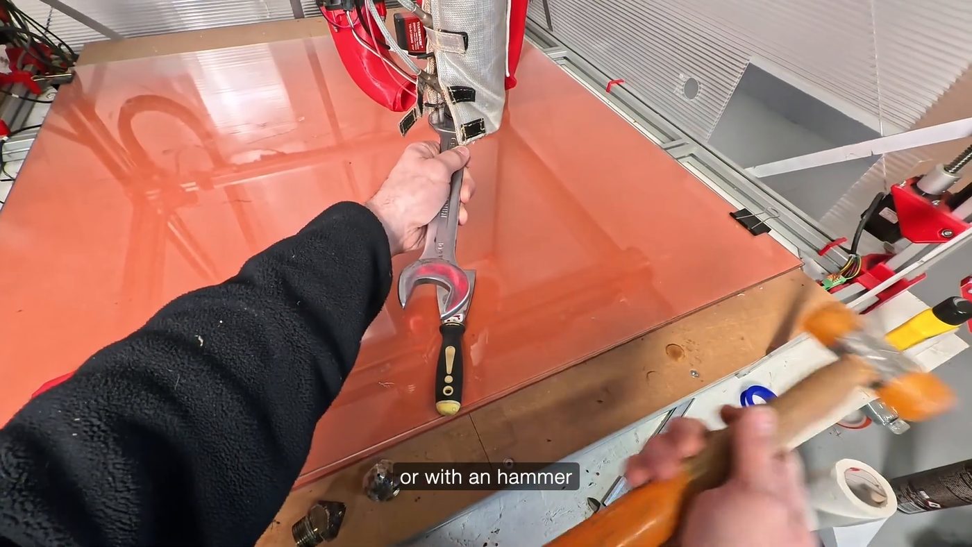 A hand holding an open-ended wrench on the G1 nozzle, with a soft mallet resting on the bench beside the machine